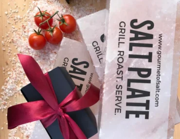 Pink Himalayan salt plates packaged with a black box wrapped in a deep red ribbon and cherry tomatoes, surrounded by loose salt crystals.